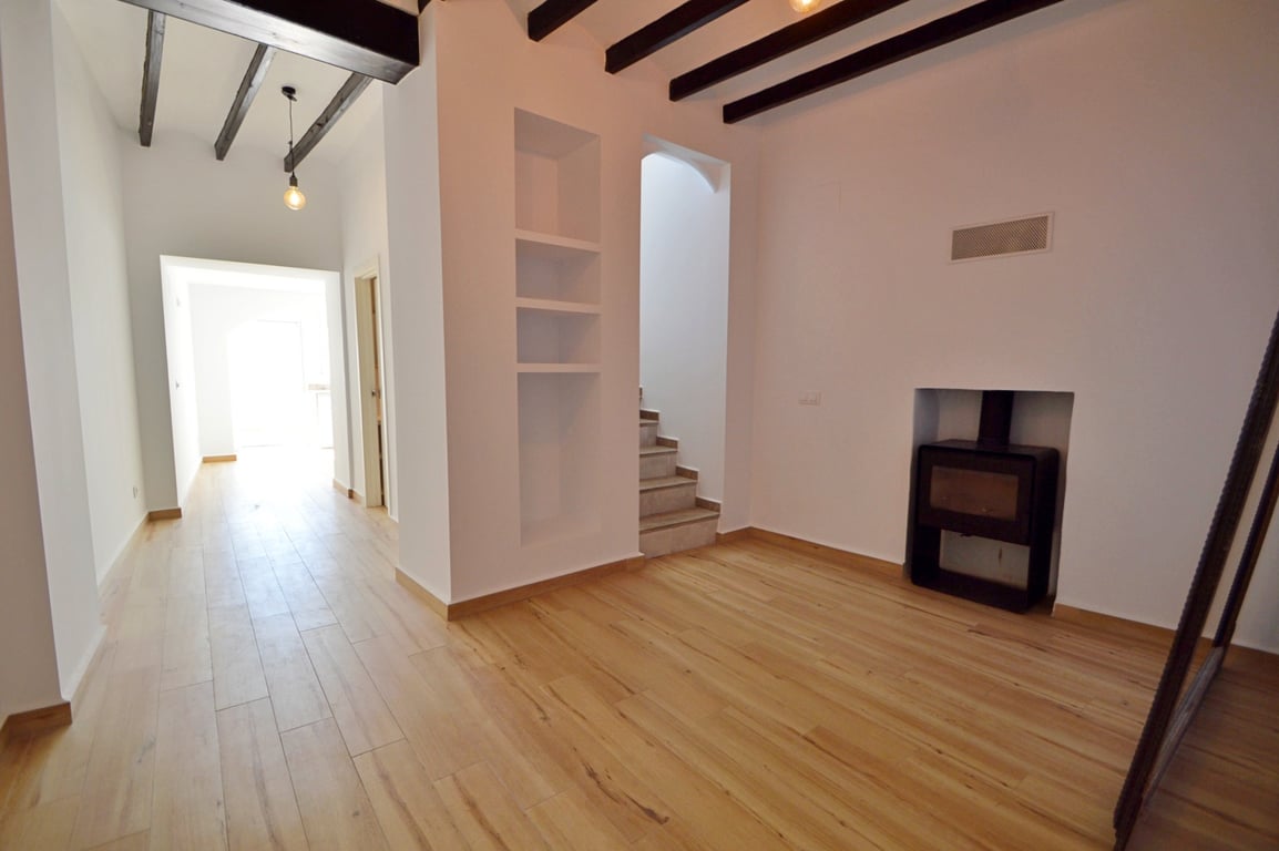 Living area featuring dark exposed ceiling beams, light wood flooring, and a built-in wood-burning stove. A recessed wall shelf and tiled staircase are visible.