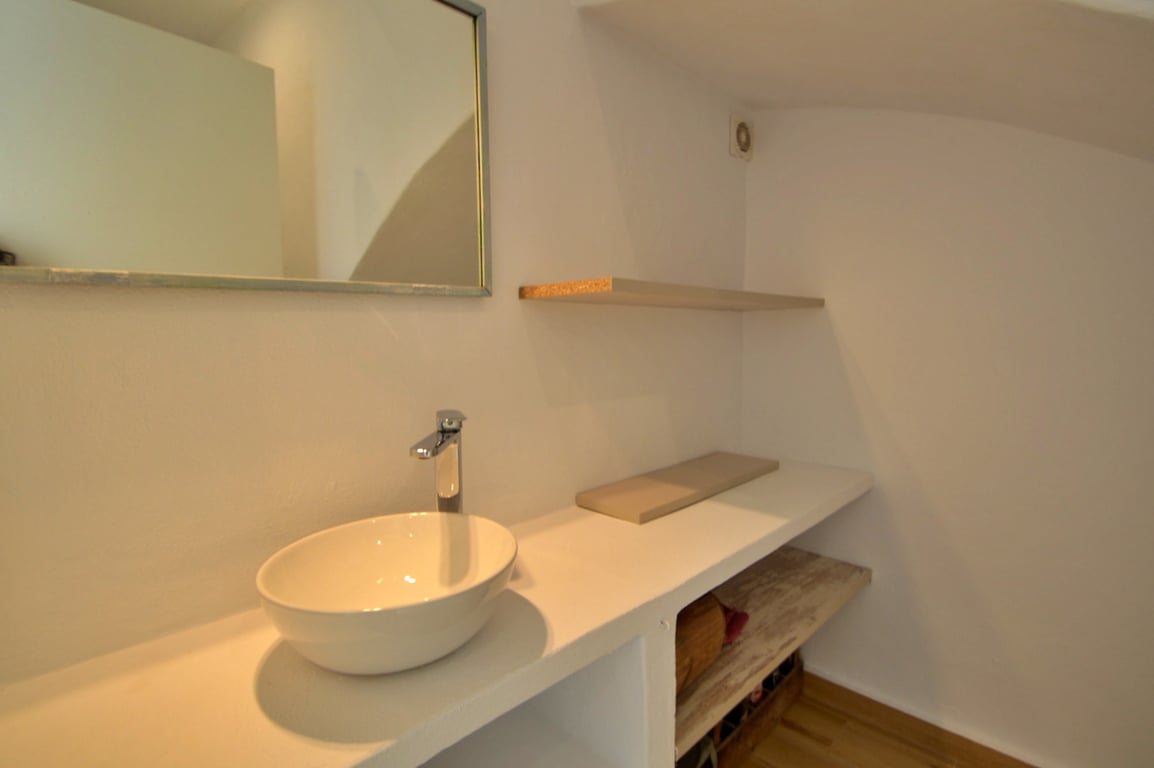 Minimalist bathroom featuring a white vessel sink on a built-in vanity, open shelving, and a rectangular mirror. The clean white walls contrast with warm wood accents.