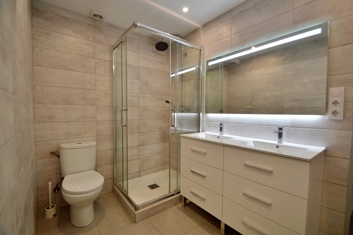 Modern bathroom featuring a double vanity with white cabinetry, a large backlit mirror, and a glass-enclosed corner shower with neutral-toned tiling.