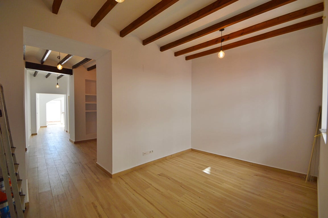 Open-plan room featuring exposed wooden ceiling beams, light wood flooring, and minimalist white walls. Pendant lighting and natural light emphasize the clean, renovated interior.