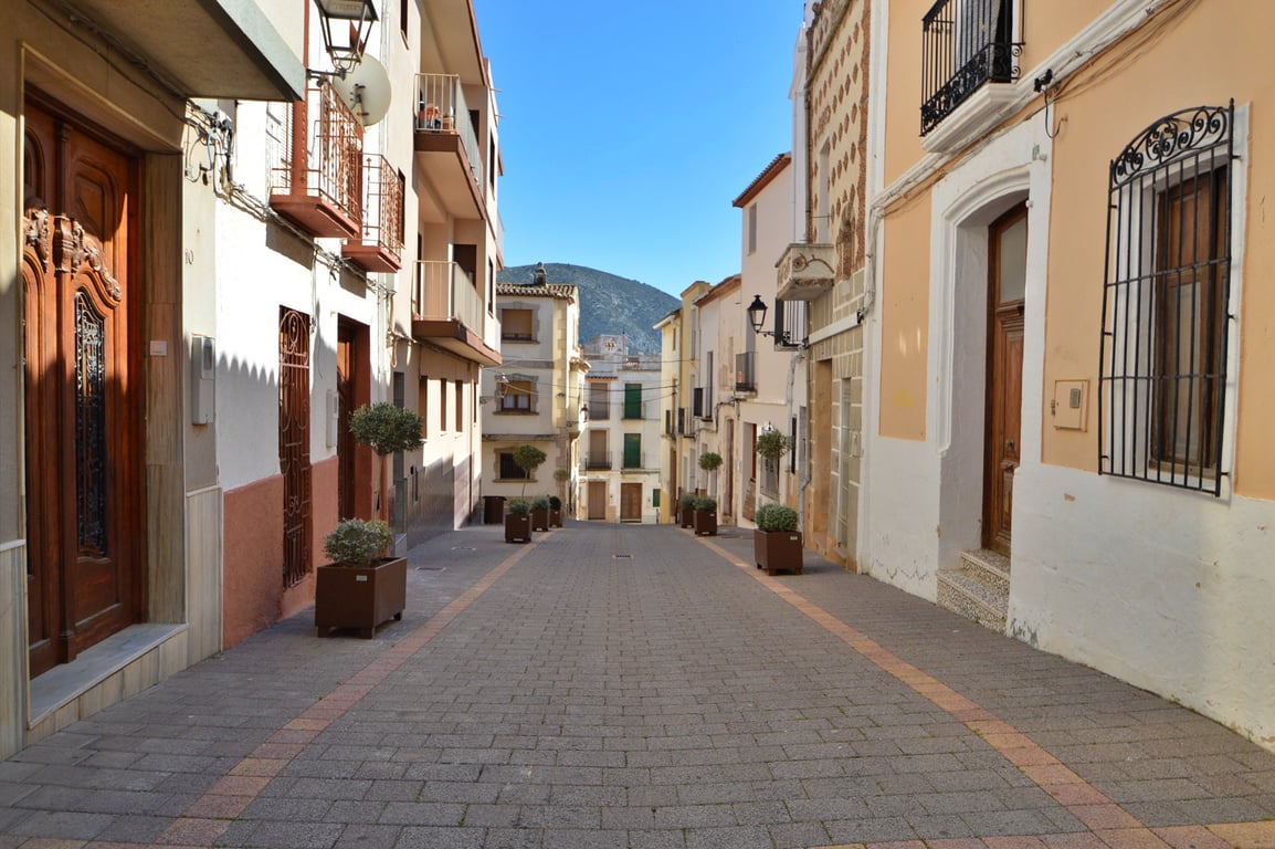 Paved pedestrian street in the village center, featuring traditional facades with wrought iron balconies and a clear view of the mountains in the distance.