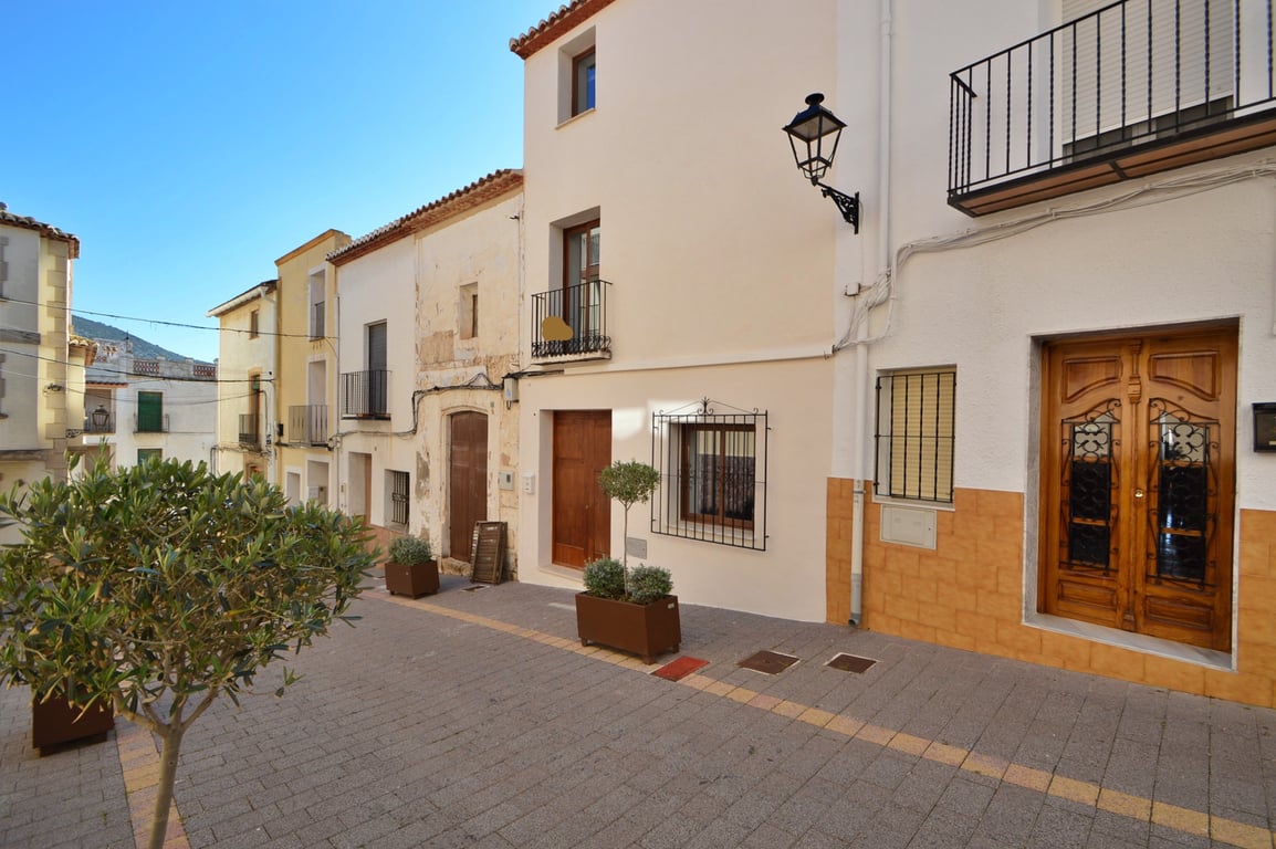 Renovated exterior facade on a paved village street featuring a wooden entrance door, wrought iron window guards, and decorative potted trees.