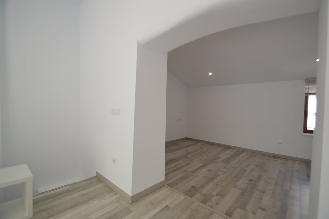 Renovated interior room featuring light wood-effect flooring, crisp white walls, and recessed ceiling lighting. The space includes a wide opening and a window with a dark frame.