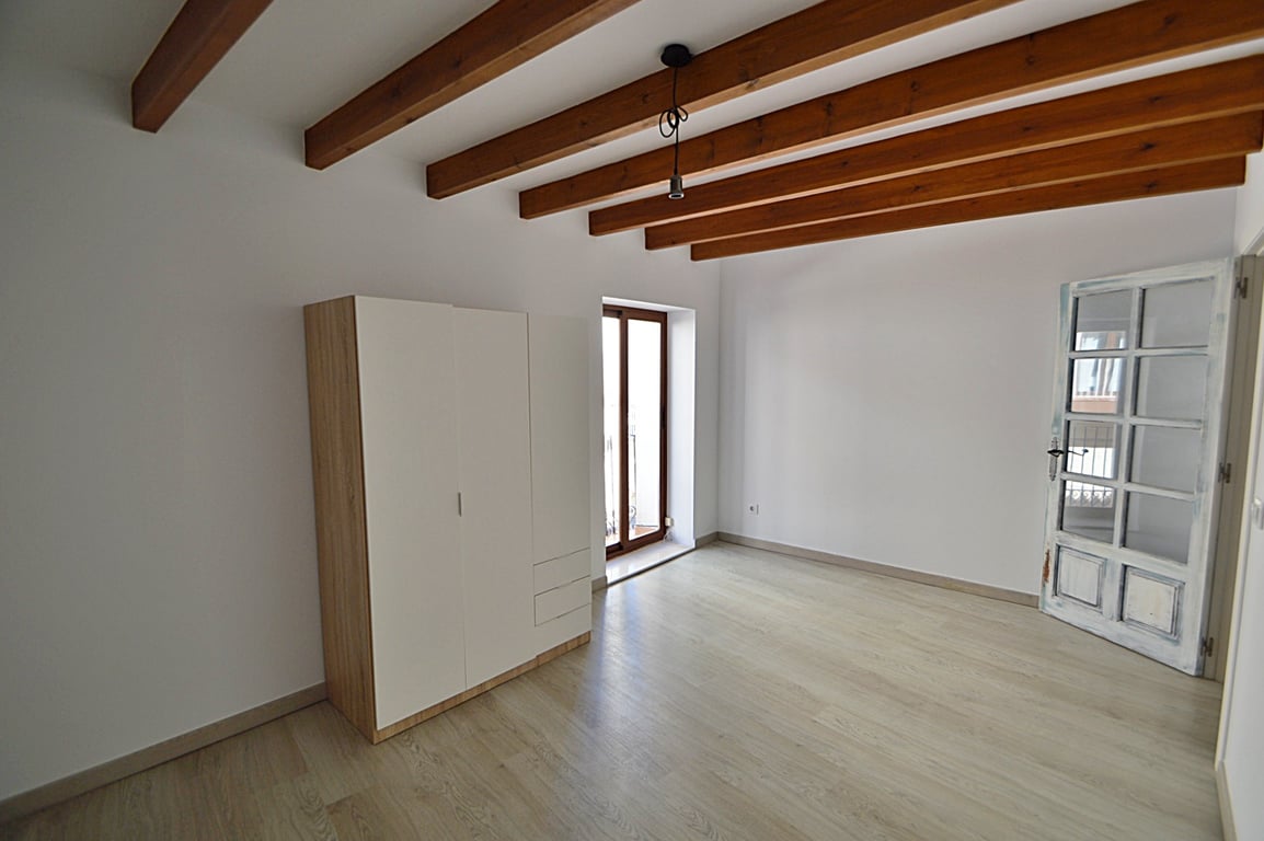 Renovated room featuring original wooden ceiling beams, light wood flooring, and balcony access. It includes a modern wardrobe and a handcrafted glazed wooden door.