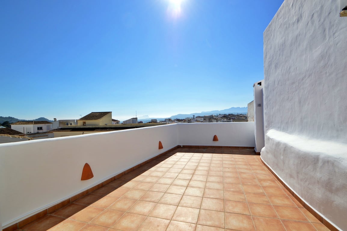 Rooftop terrace featuring terracotta tiling and white masonry walls, providing panoramic views of the surrounding town and distant mountain ranges.