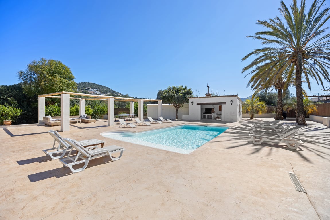 A 5x10m swimming pool surrounded by a spacious paved terrace, featuring a fully equipped outdoor kitchen, wooden pergola, and mature palm trees.