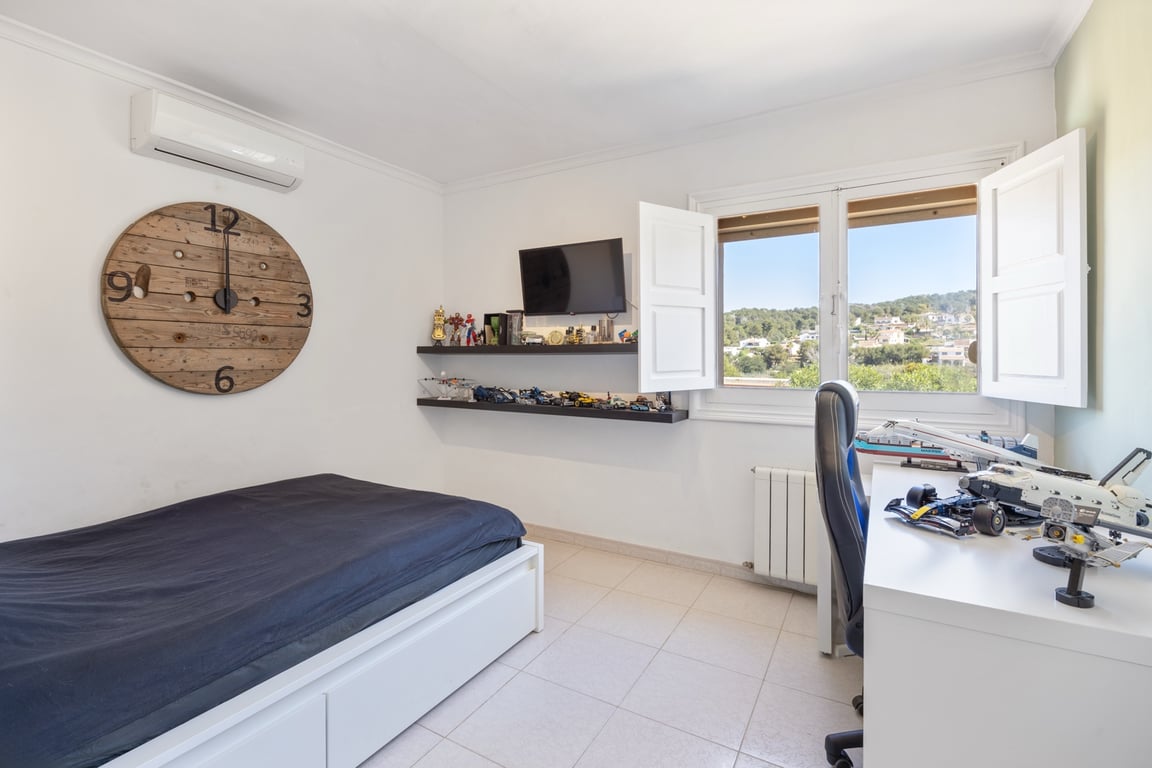 Bright bedroom featuring split-unit air conditioning, a dedicated workspace, and a window with white shutters overlooking the green hills.