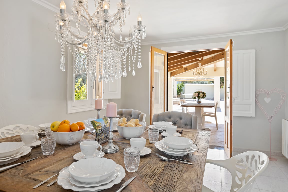 Bright dining area featuring a rustic wooden table and crystal chandelier, with double doors opening onto the covered naya and outdoor terrace.