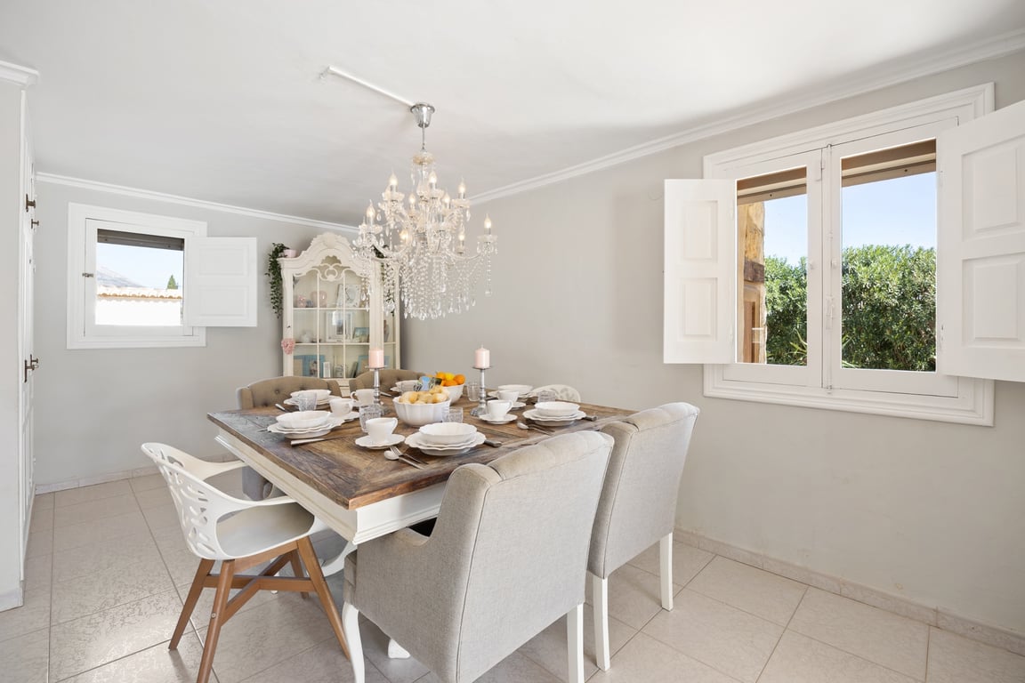 Bright dining area featuring a rustic wooden table, upholstered seating, and a crystal chandelier. White-shuttered windows provide natural light and views of the greenery.