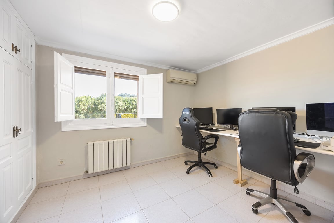 Bright home office featuring built-in wardrobes, air conditioning, and a radiator. The window with traditional shutters provides natural light and views of the surrounding greenery.