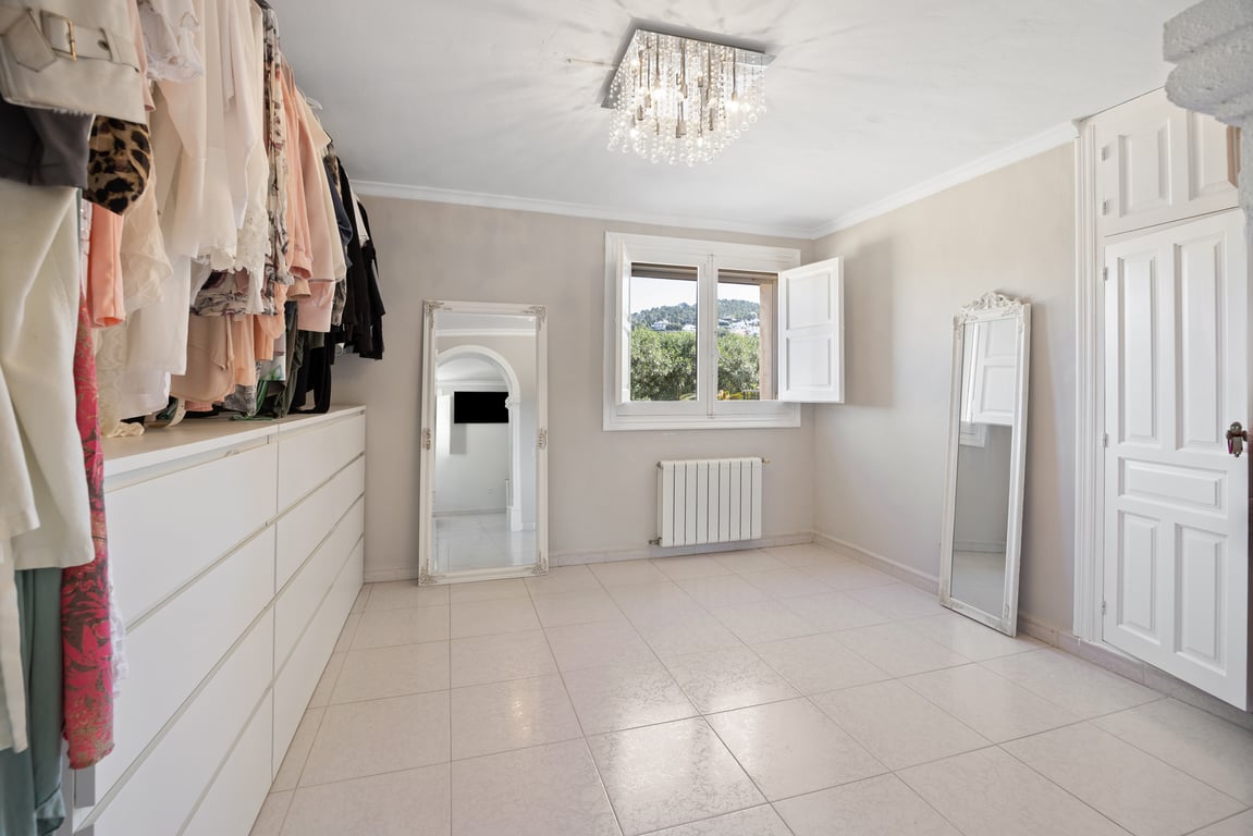 Bright walk-in closet featuring a long white chest of drawers, full-length ornate mirrors, and a crystal chandelier. The space includes central heating and garden views.