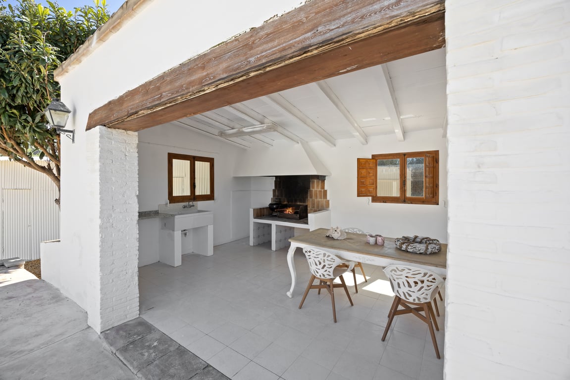 Covered outdoor kitchen featuring a built-in barbecue, stone sink, and dining area set under a white beamed ceiling with a rustic wooden lintel.