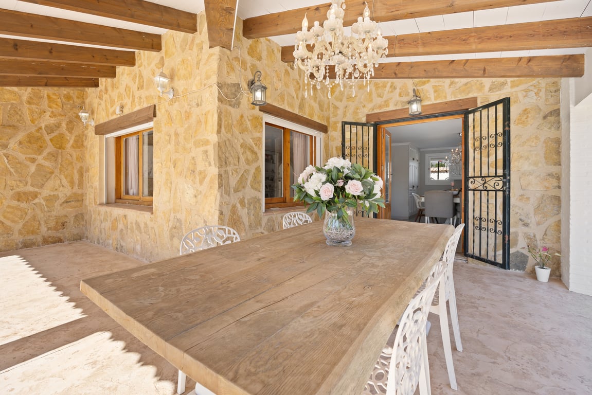Covered terrace featuring natural stone walls, exposed wooden beams, and a large dining table illuminated by a classic chandelier.
