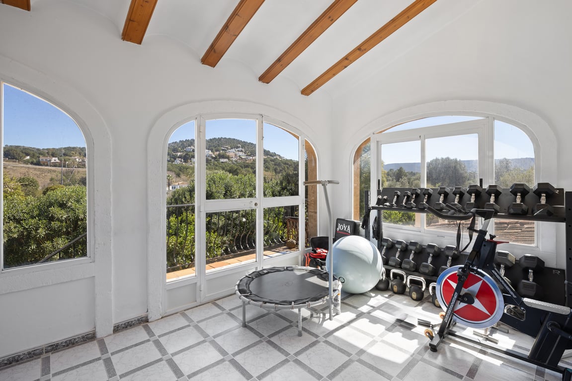 Enclosed naya converted into a home gym, featuring traditional wooden beams, tiled flooring, and arched windows with panoramic hillside views.