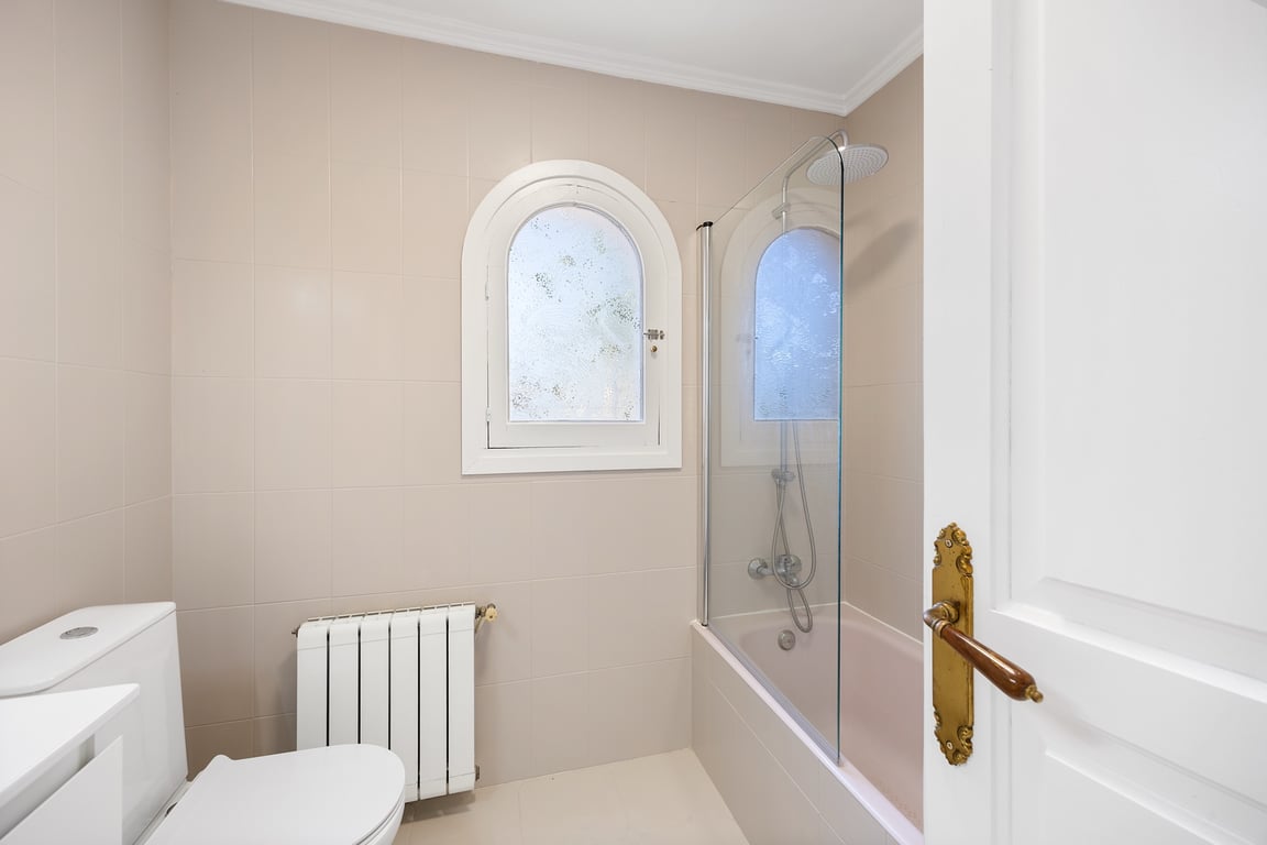 Functional bathroom featuring a bathtub with a glass screen and a distinctive arched window. It includes radiator heating and neutral-toned tiling.