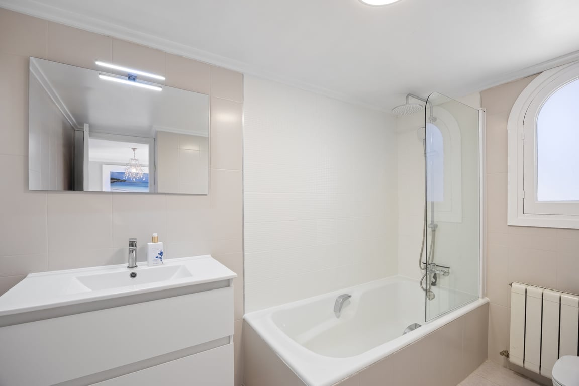 Modern bathroom featuring a white vanity, large mirror with integrated lighting, and a bathtub with a glass screen. An arched window provides natural light against neutral tiling.