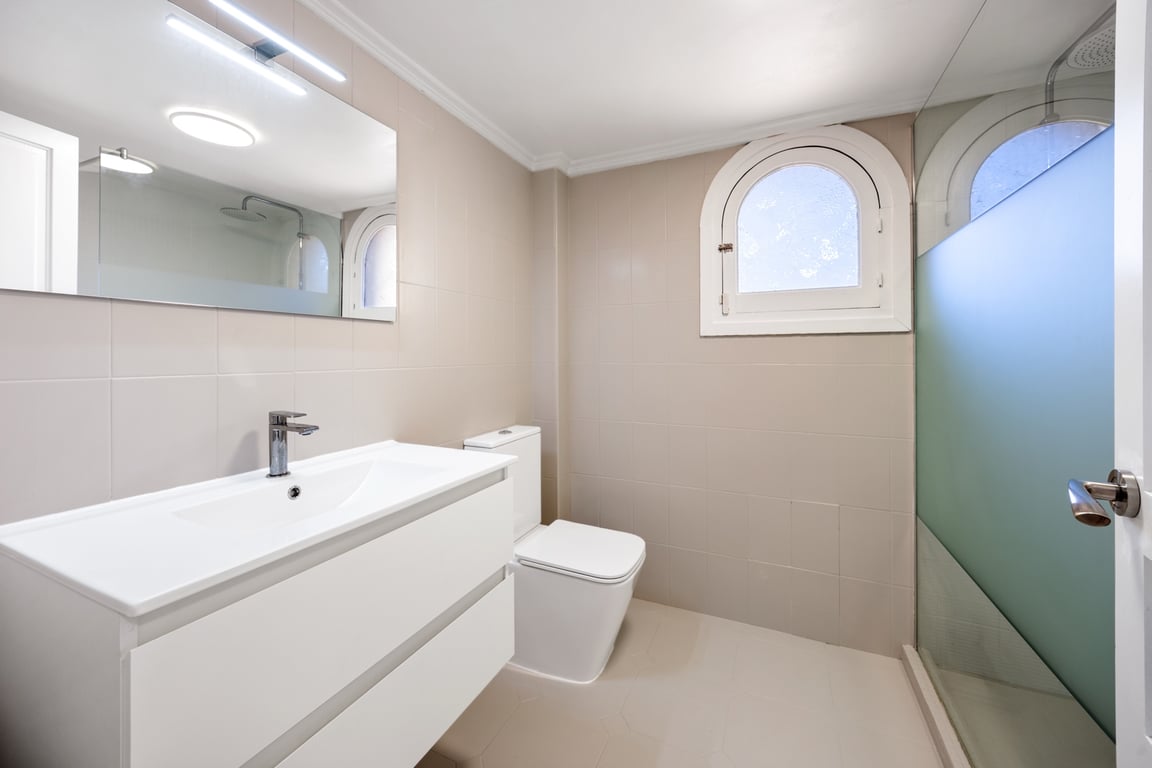 Modern bathroom featuring a white vanity, walk-in shower with frosted glass, and a traditional arched window. Neutral tiling and contemporary LED lighting create a bright space.