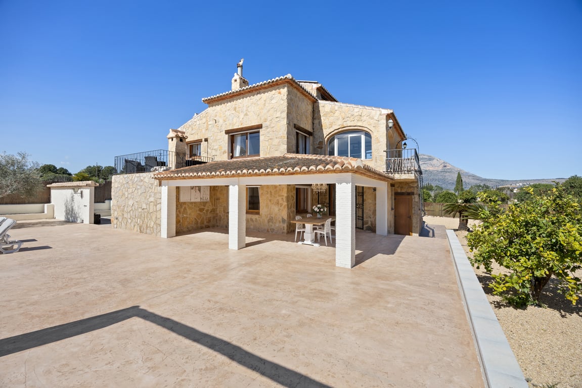 Natural stone facade featuring a covered naya with white pillars, a spacious paved terrace, and clear views of the Montgó mountain in the background.