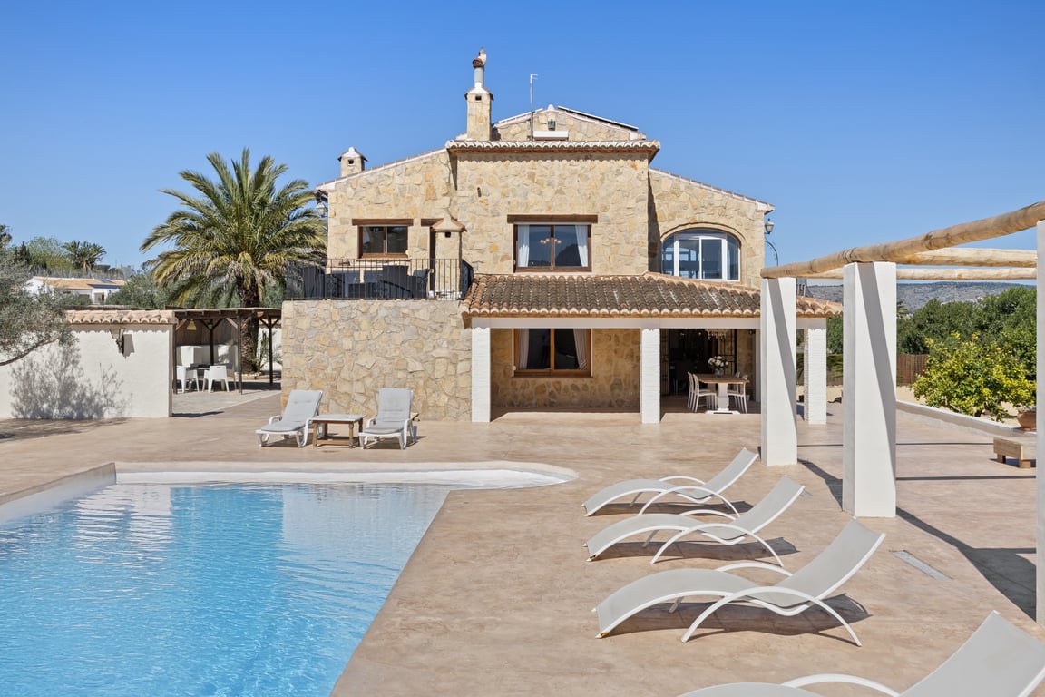 Natural stone facade featuring a covered porch, expansive tiled terrace, and swimming pool. The villa showcases its two-level architecture and sun-drenched lounge areas.