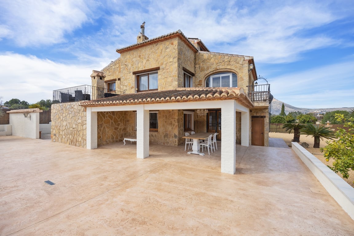 Natural stone facade featuring a covered porch with white pillars, upper-level terraces, and a spacious paved outdoor area under a clear sky.