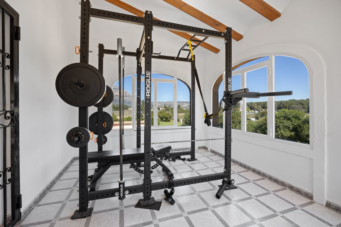 Private gym featuring professional Rogue equipment, set in a bright space with traditional wooden beams and large windows overlooking the Mediterranean landscape.