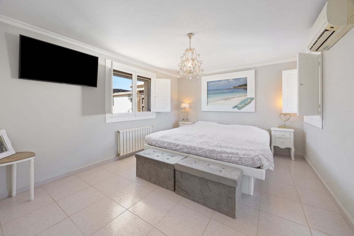 Spacious bedroom featuring white shutters, a wall-mounted TV, and split-system air conditioning. The room includes a large bed with upholstered benches.