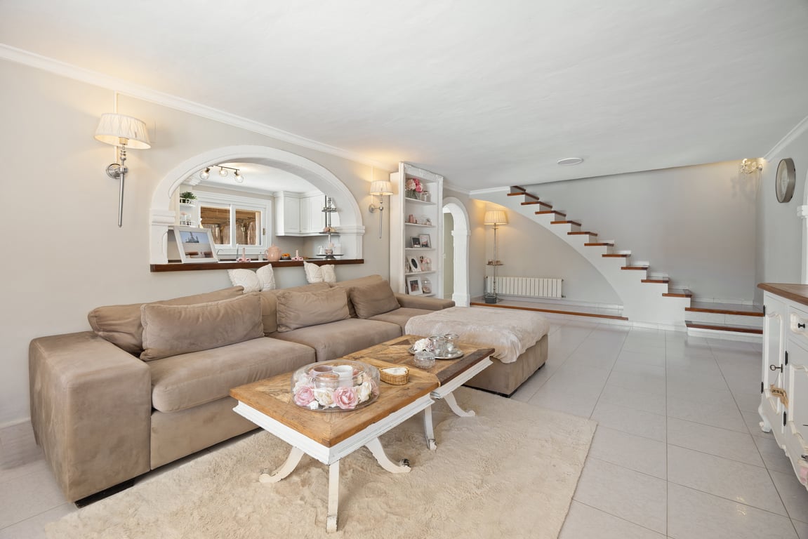 Spacious living area featuring a large sectional sofa, tiled flooring, and a decorative archway to the kitchen. A staircase with wooden treads leads to the upper level.