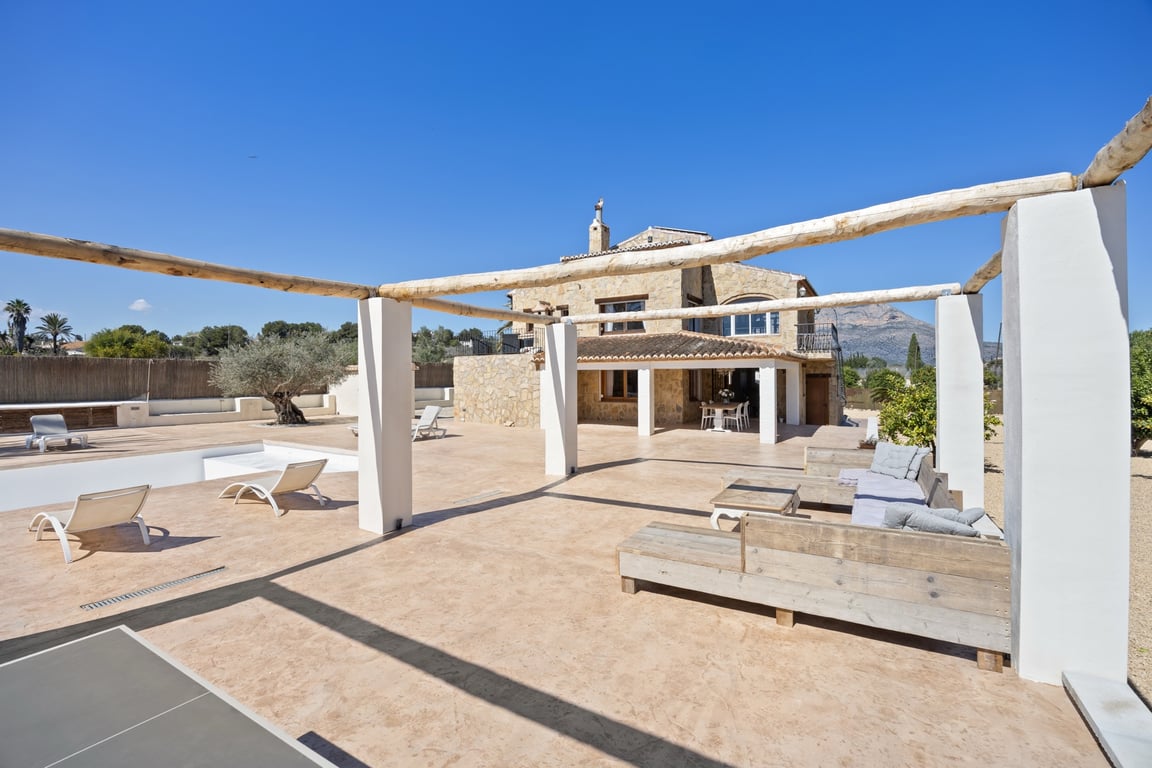 Spacious outdoor terrace featuring a wooden beam pergola, lounge seating, and views of the stone-clad villa against the Montgó mountain backdrop.