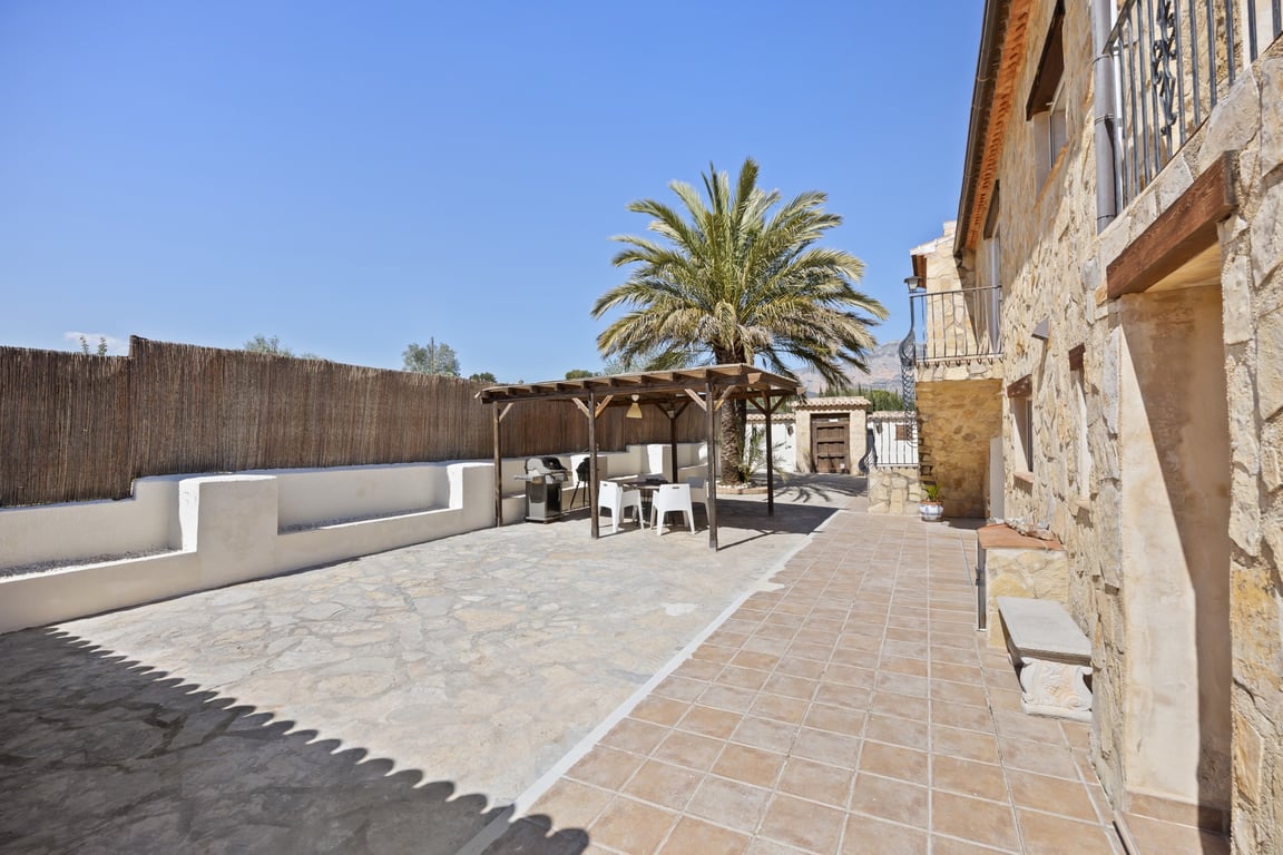 Spacious outdoor terrace featuring stone paving, a wooden pergola with a dining area and BBQ, set against a traditional stone-clad villa facade.