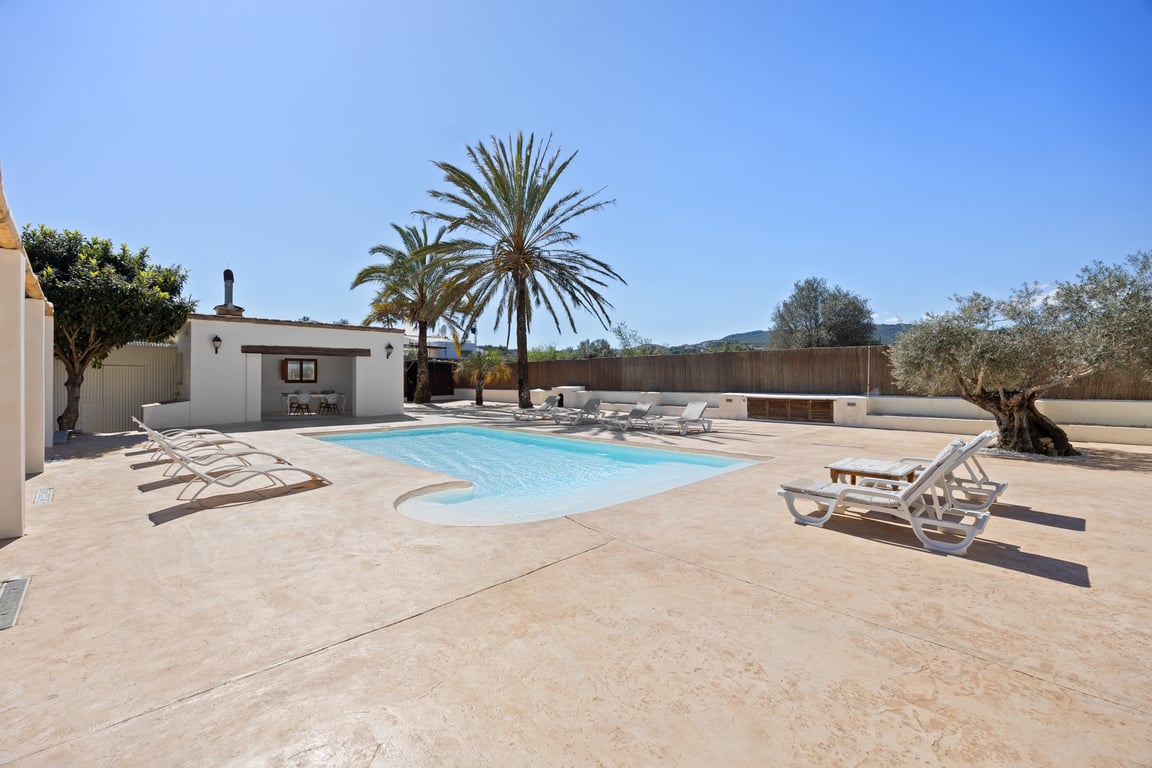 Spacious pool terrace with a curved entry basin, featuring a large sunbathing area, Mediterranean trees, and a detached outdoor kitchen building.