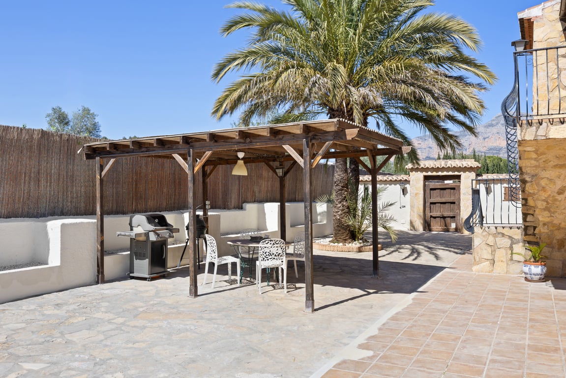 Wooden pergola shading an outdoor dining area with a gas barbecue, set on a stone-paved terrace featuring a mature palm tree and mountain views.