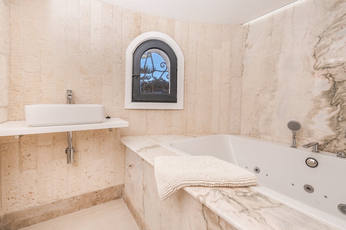 Bathroom featuring a marble-encased jetted tub, a vessel sink, and textured limestone walls with a traditional arched window.