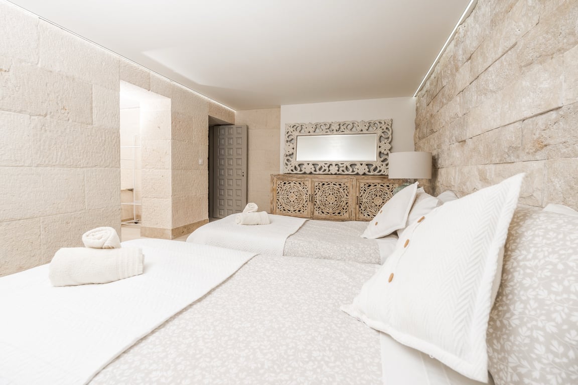 Bedroom featuring natural limestone block walls, twin beds, and ornate carved wooden furniture. Indirect LED strip lighting highlights the stone textures.