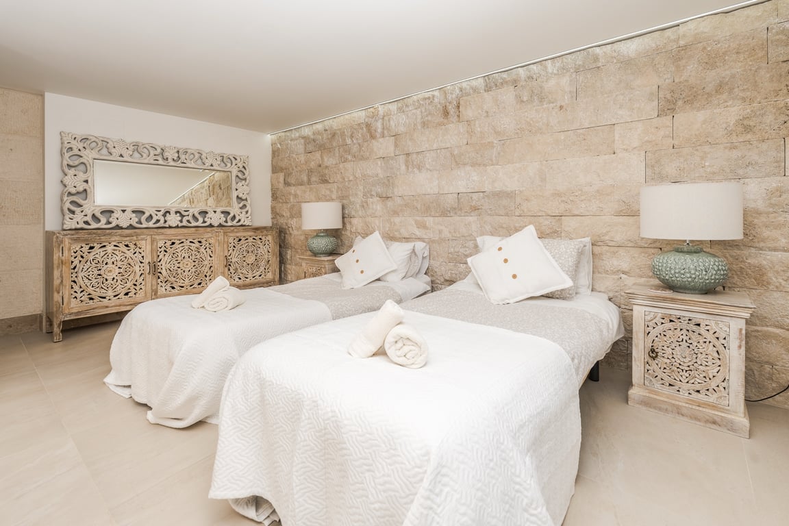Bedroom featuring twin beds set against a textured natural stone wall. Includes hand-carved wooden furniture, ceramic lamps, and integrated recessed ceiling lighting.