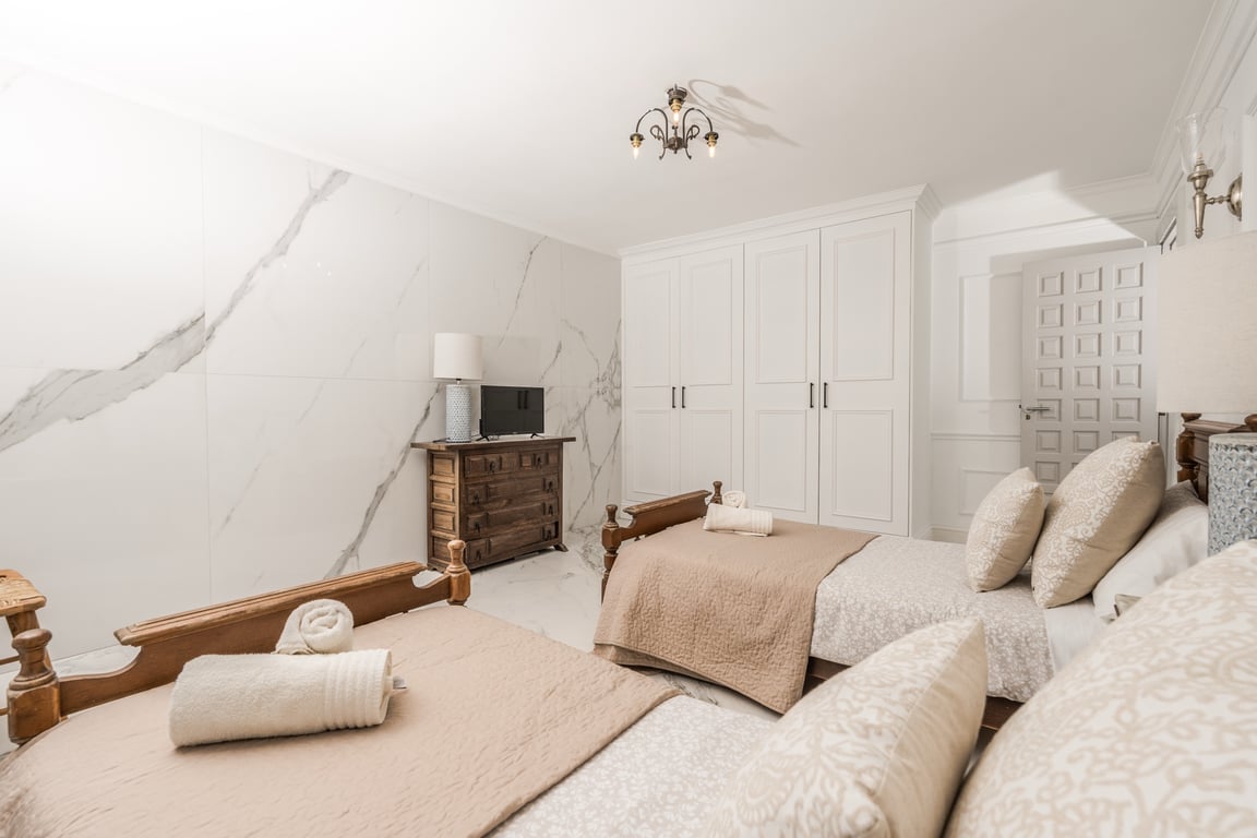 Bedroom featuring twin wooden beds, a marble-effect accent wall, integrated white wardrobes, and a rustic wooden dresser with a television.