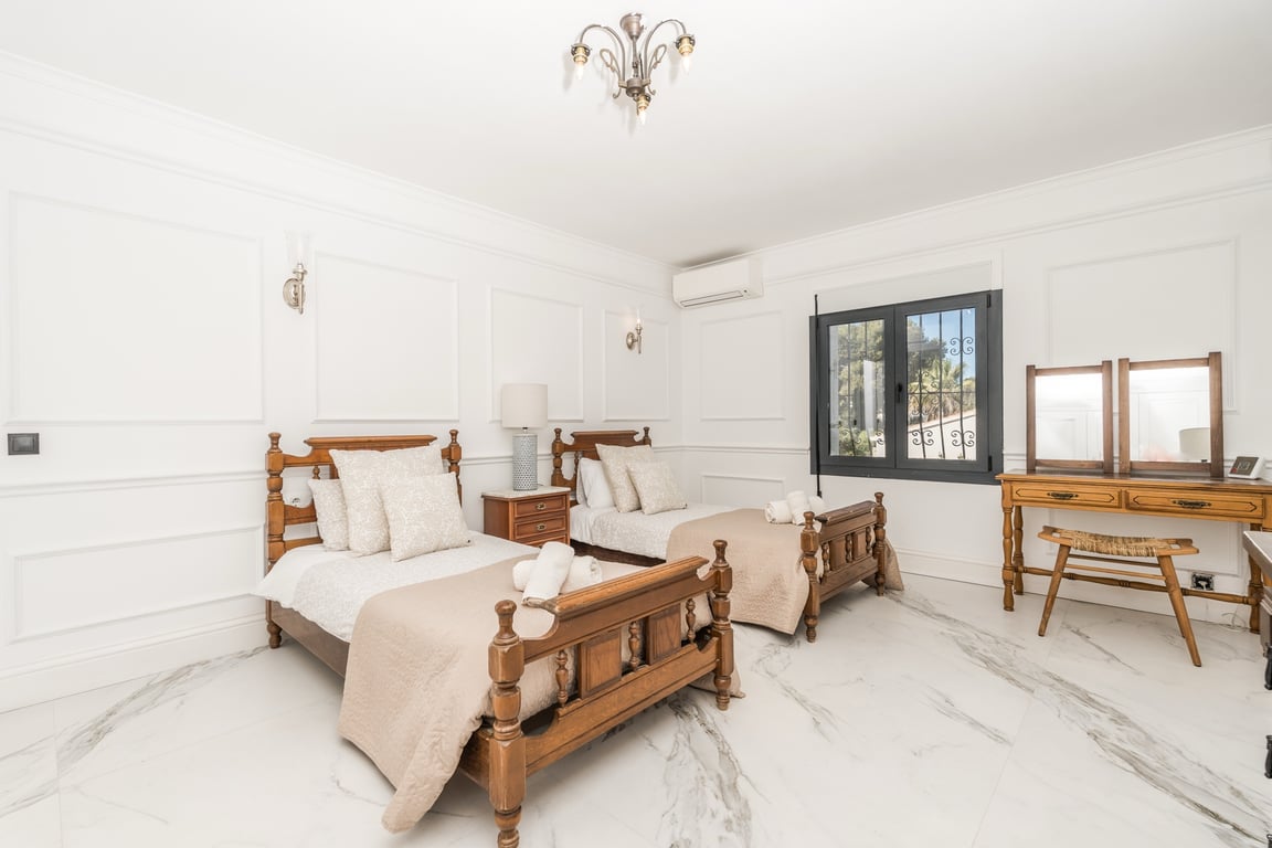 Bedroom featuring two carved wooden twin beds, marble flooring, and classic wall moldings. A wooden vanity table and air conditioning unit complete the space.