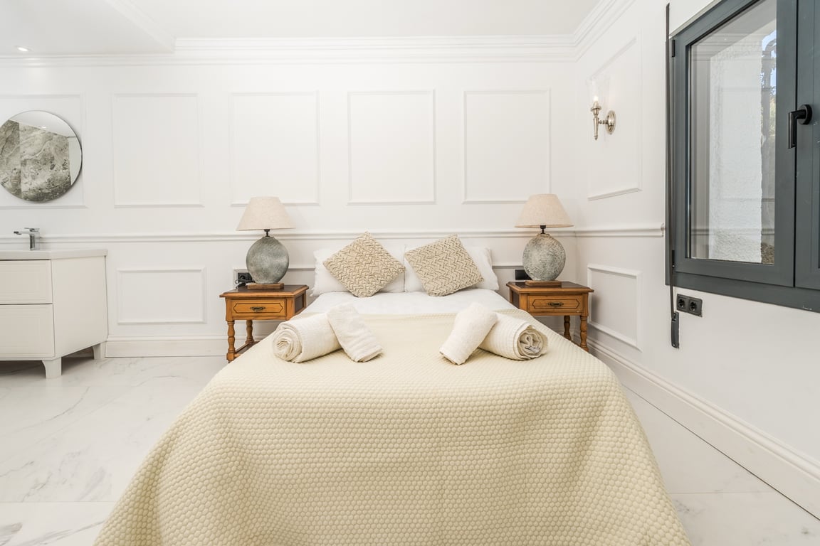 Bedroom featuring white wall paneling and marble flooring. It includes an integrated vanity area with a round mirror and classic wooden bedside tables.