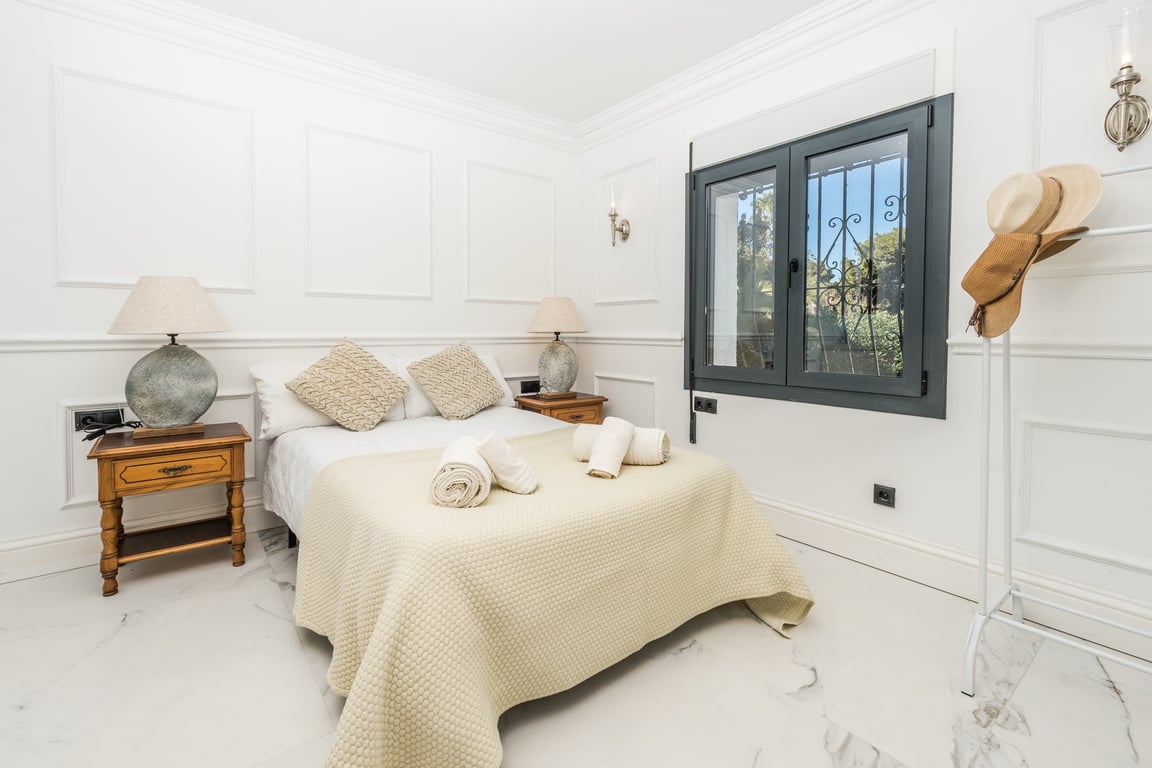 Bright bedroom featuring decorative wall moldings, white marble flooring, and dark-framed windows. Furnished with wooden bedside tables and stone-base lamps.