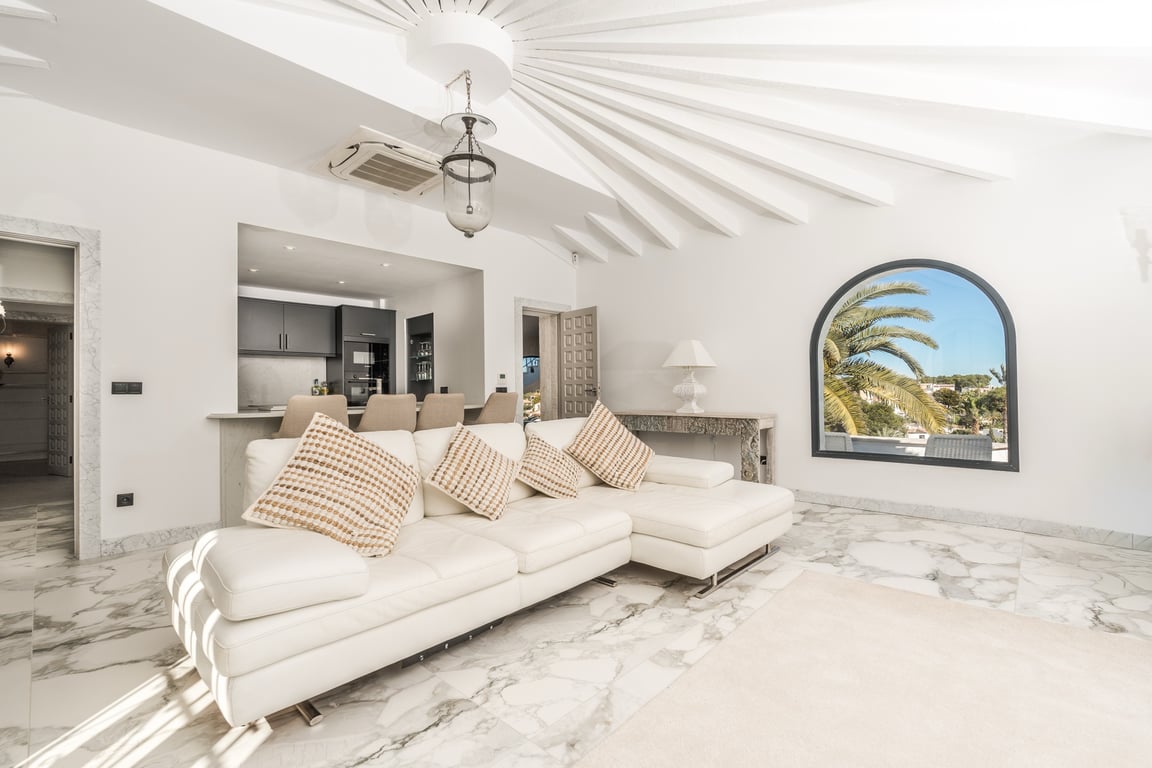 Bright living area featuring marble flooring, a distinctive radial beam ceiling, and a white leather sofa. An arched window frames palm views, connecting to the semi-open kitchen.