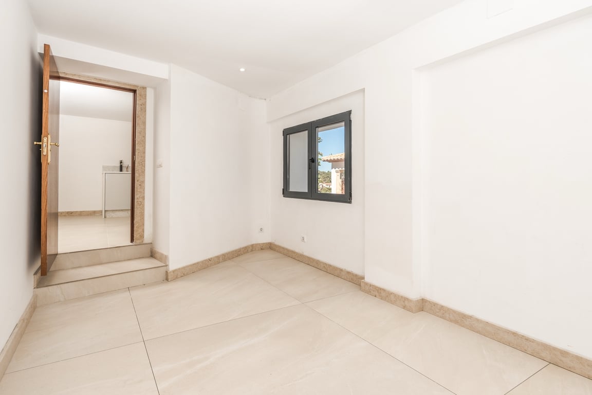 Bright room featuring large-format stone flooring, matching baseboards, and white walls. Includes a dark-framed window and a wooden door leading to an adjacent area.