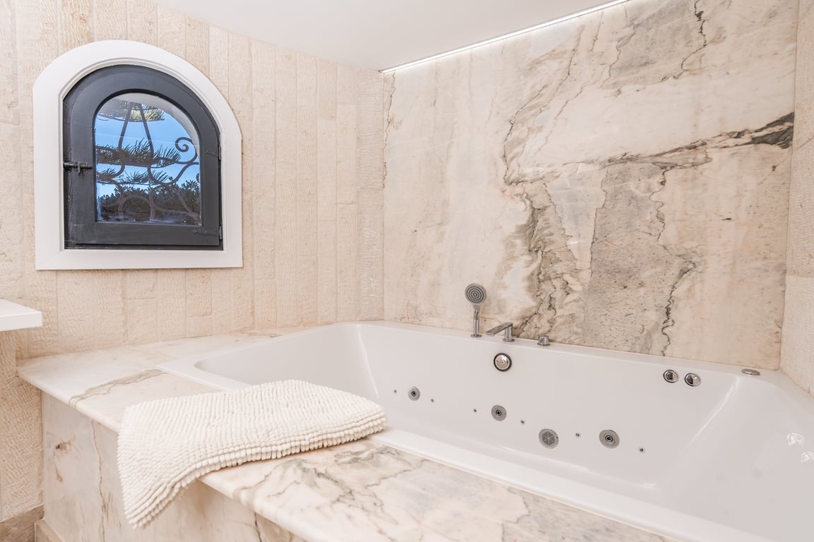Built-in whirlpool bathtub featuring elegant marble surrounds, textured stone walls, and a traditional arched window with ironwork.