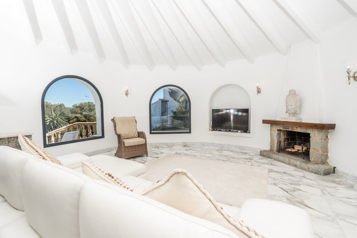 Circular living room featuring a vaulted ceiling with white radial beams, marble flooring, and a stone fireplace. Arched windows provide natural light and garden views.