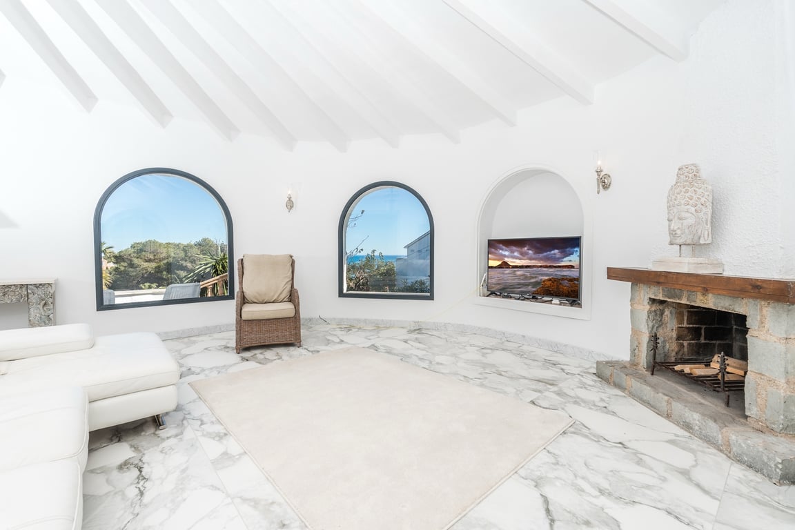 Circular living room featuring white beamed ceilings, marble flooring, and a rustic stone fireplace. Arched windows provide natural light and views of the Mediterranean landscape.