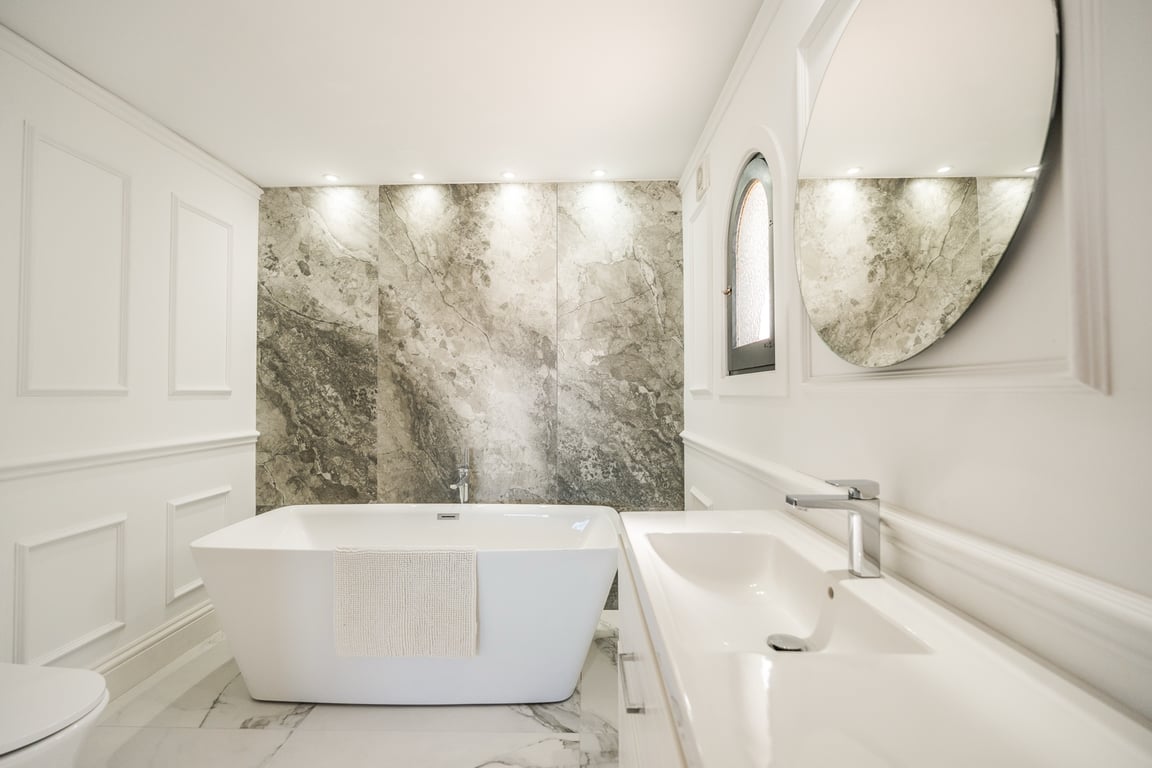Elegant bathroom featuring a freestanding bathtub, marble-effect accent wall with recessed lighting, and classic white wall paneling.
