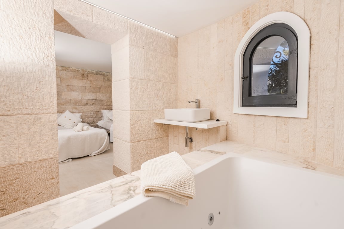 En-suite bathroom featuring natural stone walls, a marble-trimmed bathtub, and a modern vessel sink, with a view into the adjoining bedroom through a stone archway.