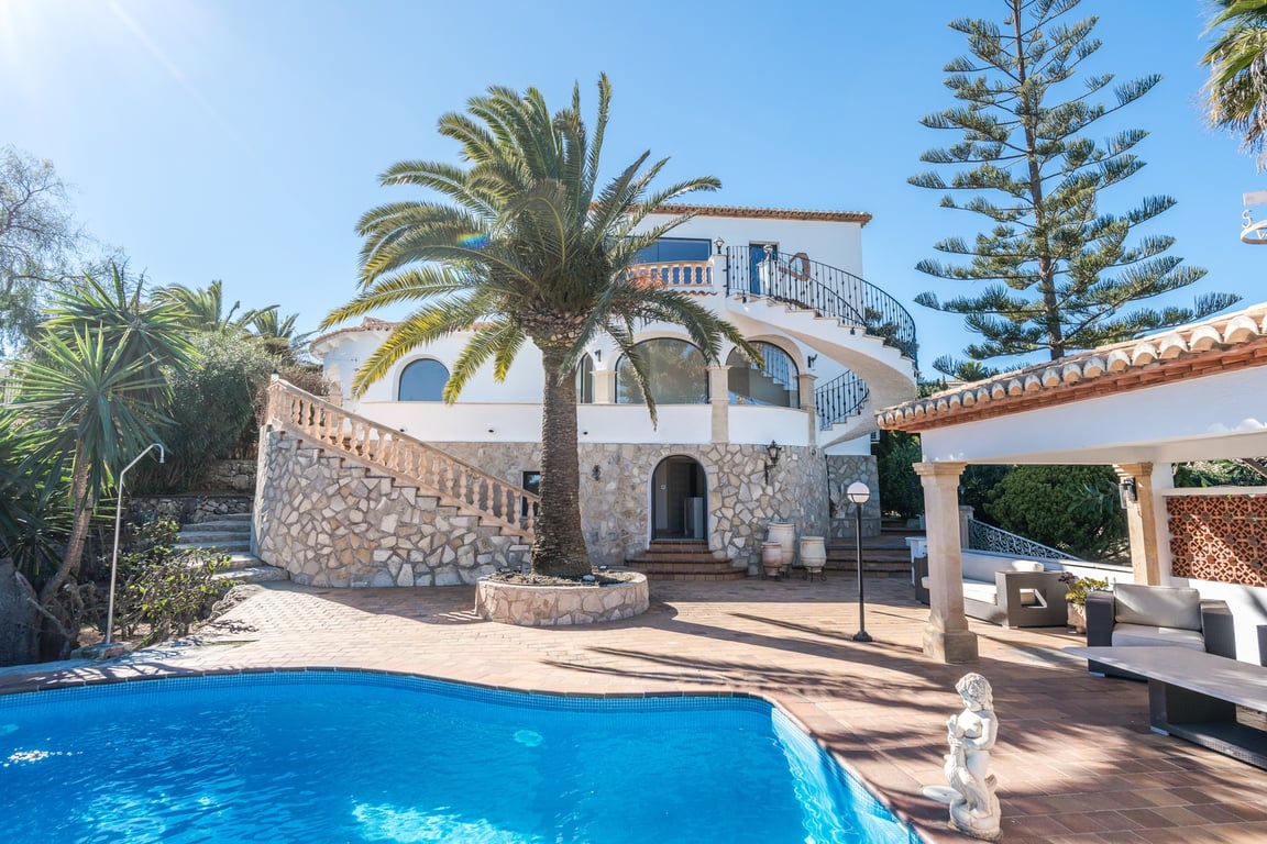 Exterior view of the villa featuring a curved swimming pool, stone-clad walls, a central palm tree, and elegant spiral staircases leading to the upper terraces.