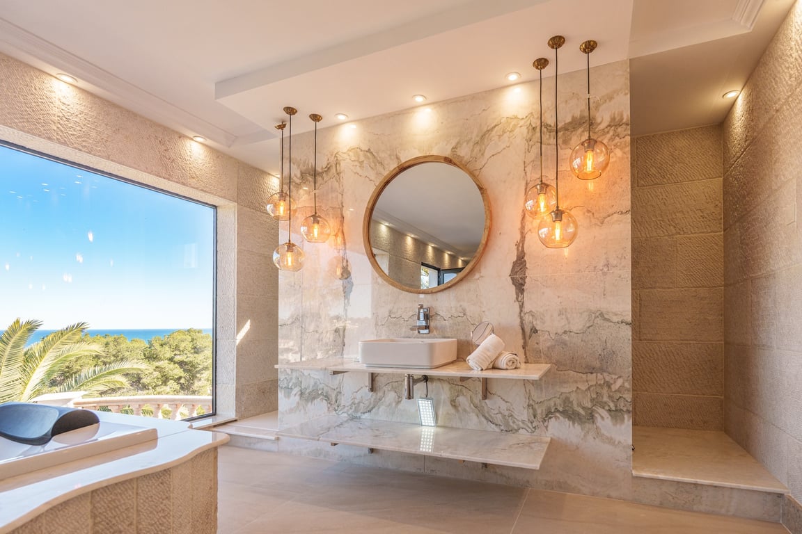Master bathroom featuring veined marble walls, a floating vanity with a vessel sink, and a circular mirror. Globe pendant lighting and a large window offer views of the Mediterranean.