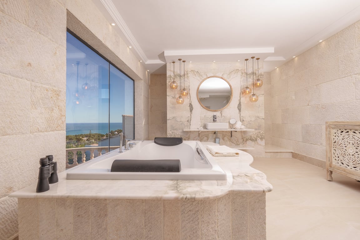 Master suite bathroom featuring limestone walls, a marble-encased bathtub with sea views, and a designer vanity area with elegant glass pendant lighting.