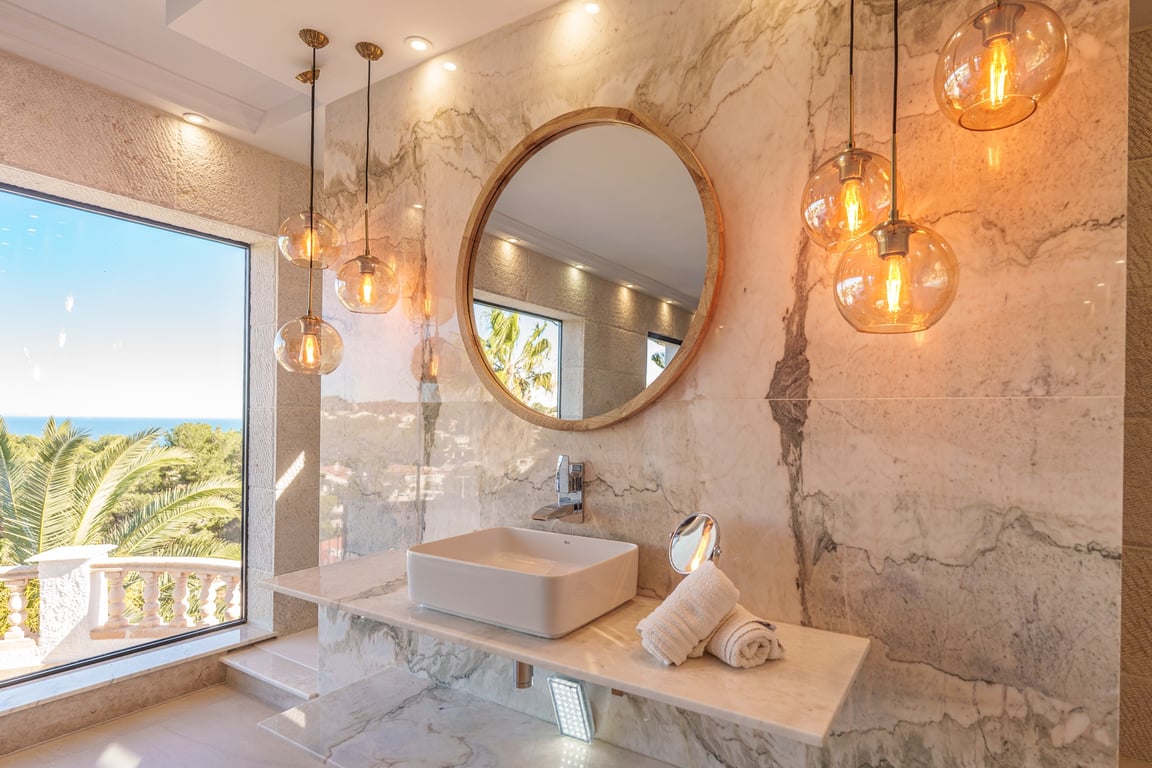 Master suite bathroom featuring marble wall cladding, a vessel sink, a round wooden mirror, and amber glass pendants with views of the Mediterranean.