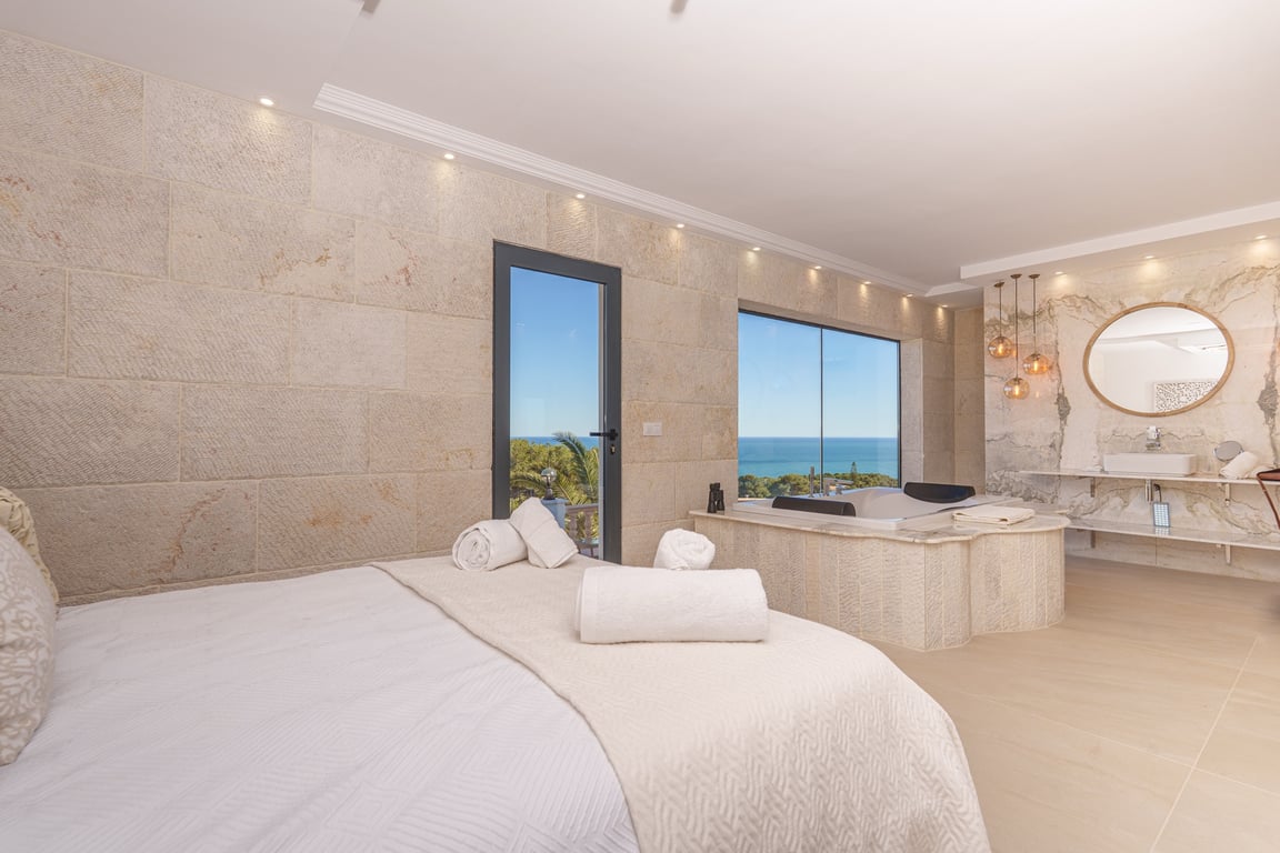Master suite featuring natural stone walls, an integrated jacuzzi by the sea-view window, and an open-concept bathroom with marble accents.